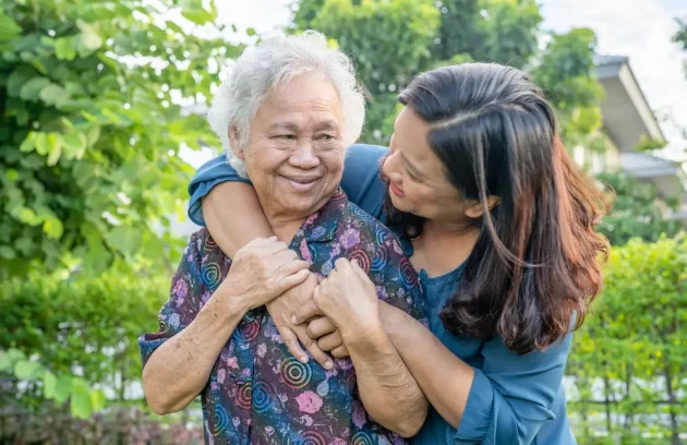 Younger woman hugs a smiling older woman from behind outdoors in a sunny garden
